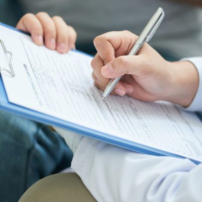 Crop shot of anonymous doctor in white gown writing with pen on clipboard during home visit